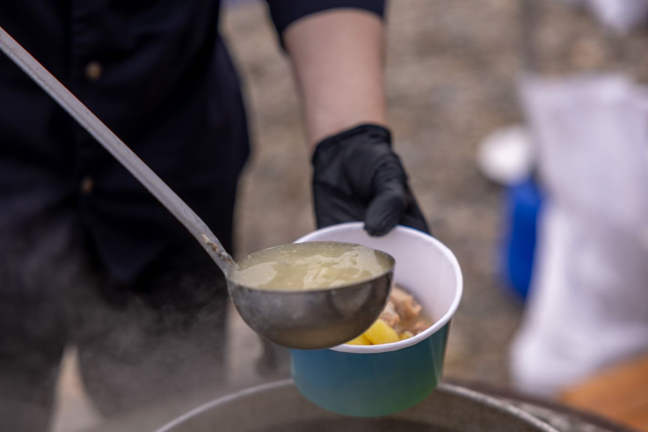 A person serves steaming hot soup from a pot into a bowl using a ladle outdoors.