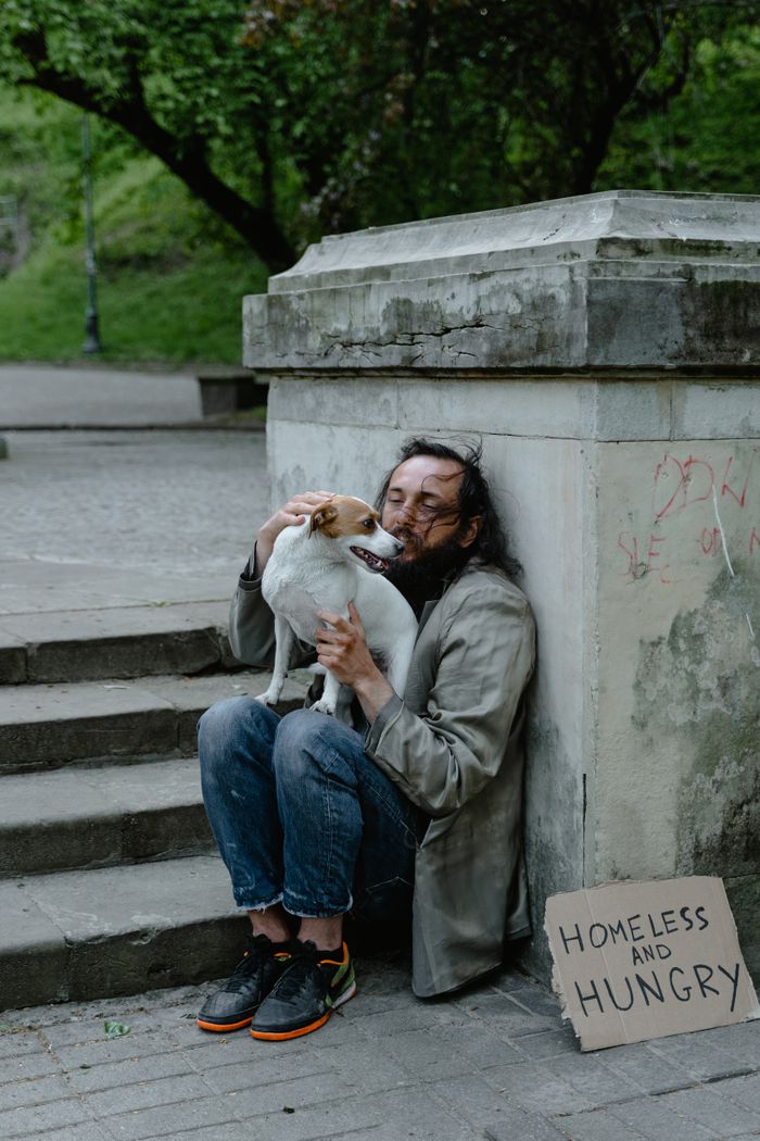A homeless man embraces his dog next to a sign reading 'Homeless and Hungry' on the city streets.
