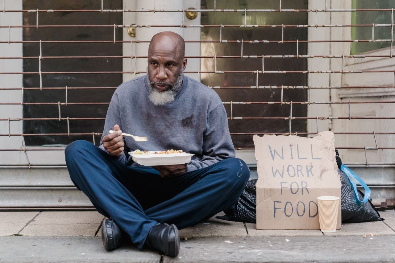 Elderly homeless man sitting on street with a sign and meal, symbolizing poverty.